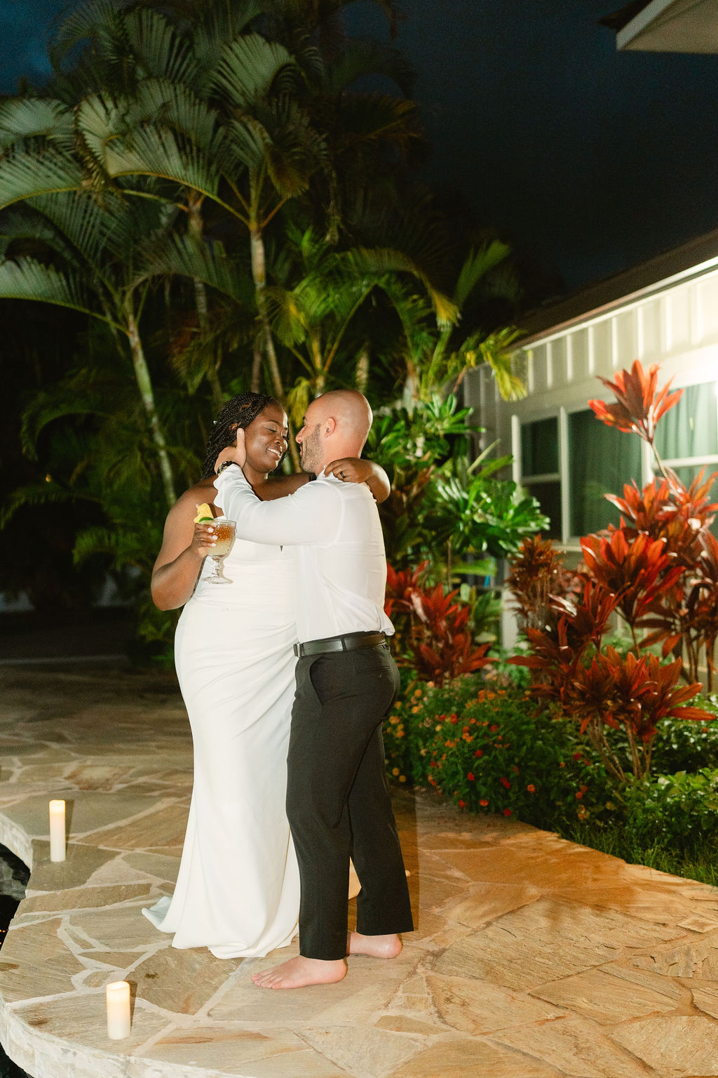 A couple stands on a stone walkway by a pool, embracing and holding a drink. Lush greenery and a building are in the background.