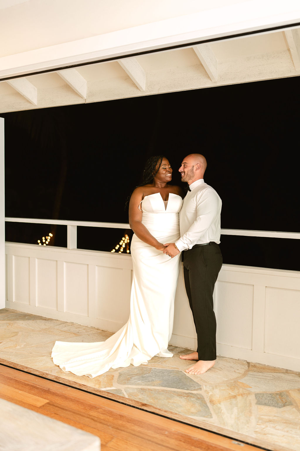 A couple, dressed in wedding attire, stands holding hands on a stone floor at night. The bride wears a white gown, and the groom is in a shirt and pants, barefoot.