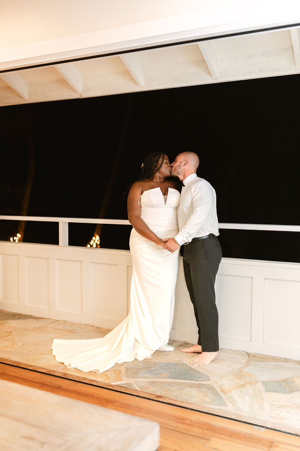 A couple, dressed in wedding attire, stands holding hands on a stone floor at night. The bride wears a white gown, and the groom is in a shirt and pants, barefoot.