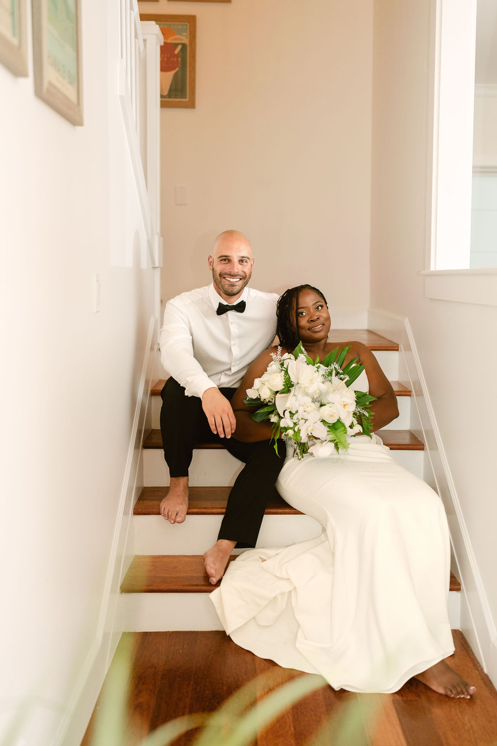 A bride and groom sit on stairs, barefoot. The groom wears a white shirt and bow tie, while the bride in a white dress holds a bouquet of white flowers. Both appear relaxed and smiling for a micro wedding in oahu