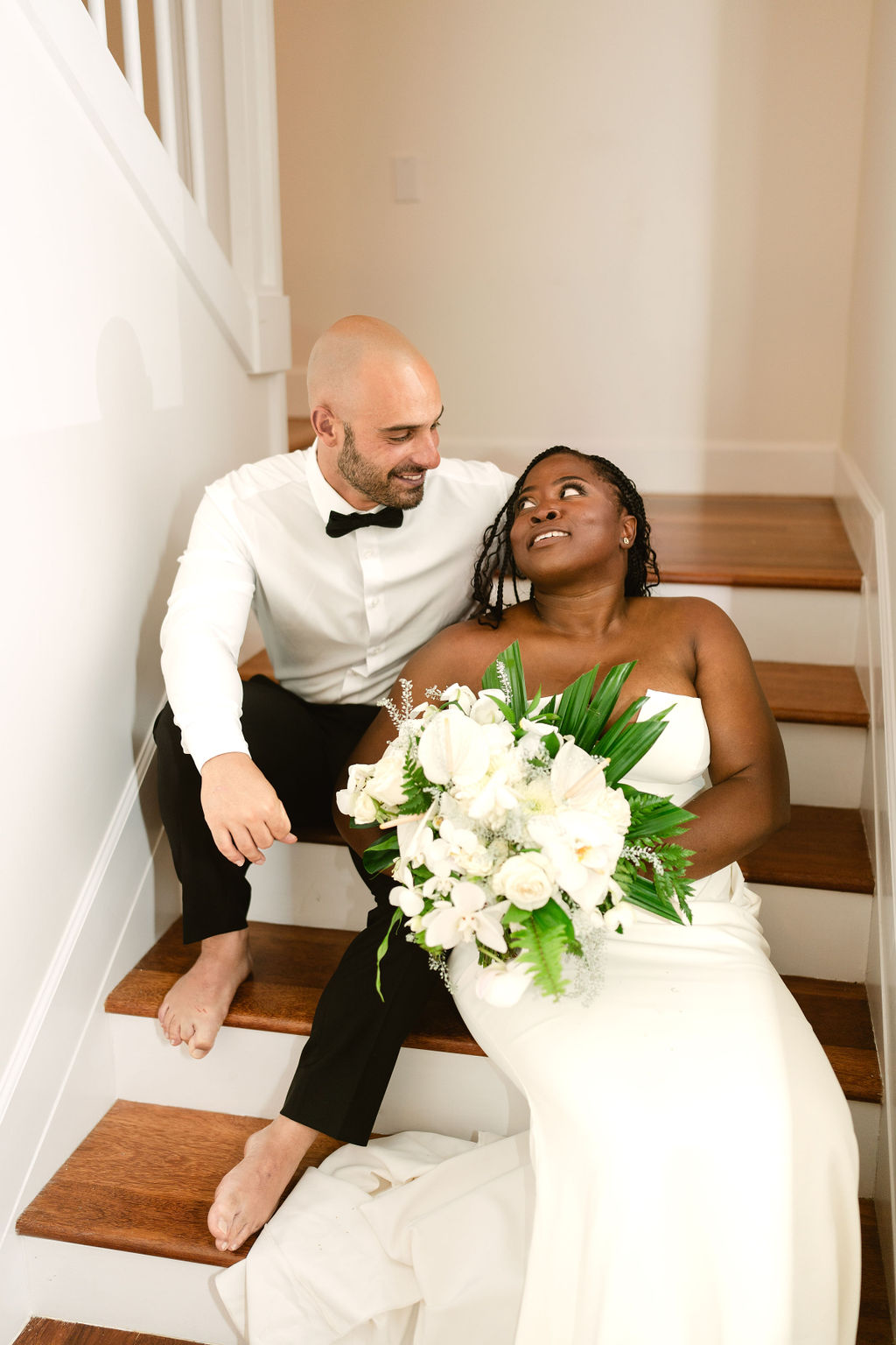 A bride and groom sit on stairs, barefoot. The groom wears a white shirt and bow tie, while the bride in a white dress holds a bouquet of white flowers. Both appear relaxed and smiling for a micro wedding in oahu