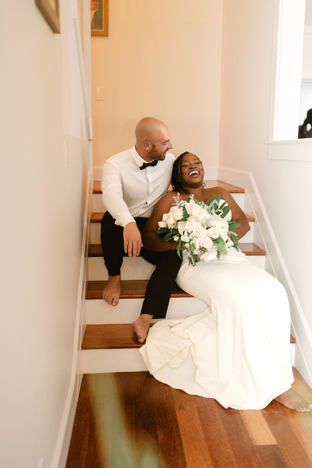 A bride and groom sit on stairs, barefoot. The groom wears a white shirt and bow tie, while the bride in a white dress holds a bouquet of white flowers. Both appear relaxed and smiling for a micro wedding in oahu