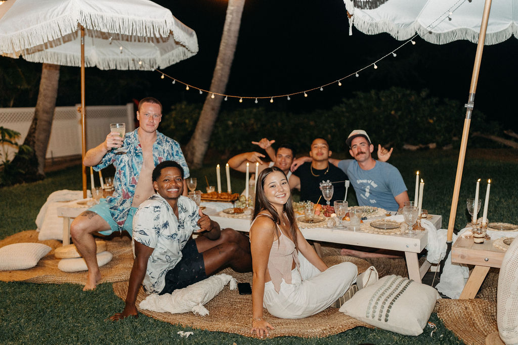 A group of six people sitting around a low outdoor dining table with food and drinks, under white umbrellas and string lights, in a nighttime setting.