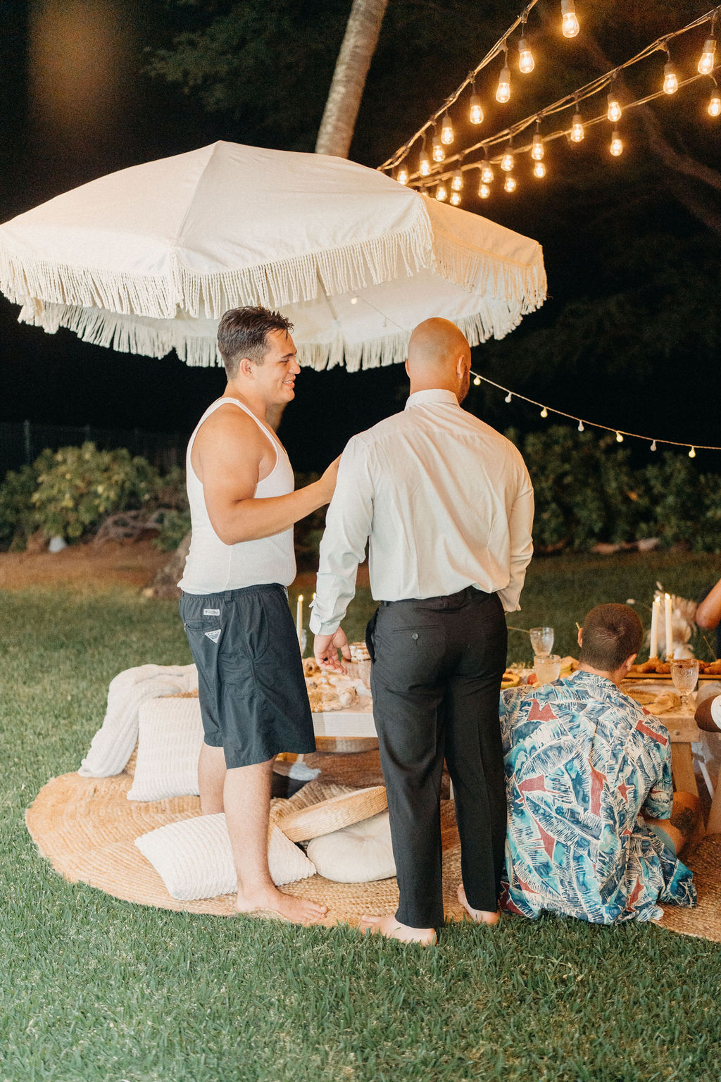 Two men stand under a fringed umbrella at an outdoor gathering with string lights. Other people sit on the ground with cushions and food nearby.