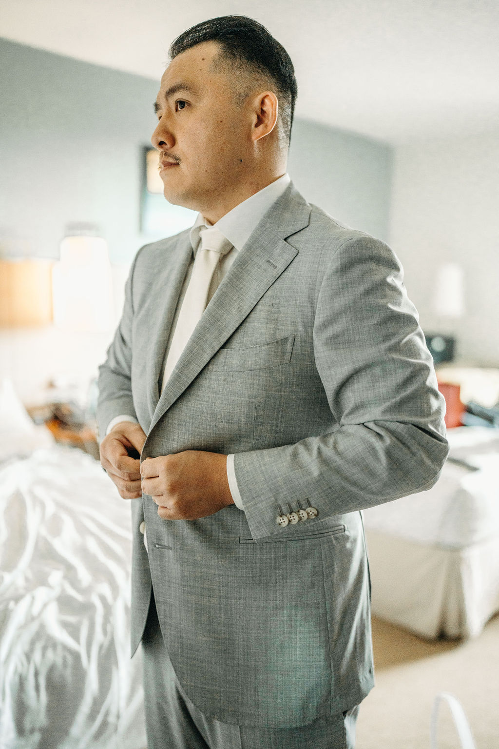 Man in a light gray suit stands smiling in a room with a bed and soft lighting.