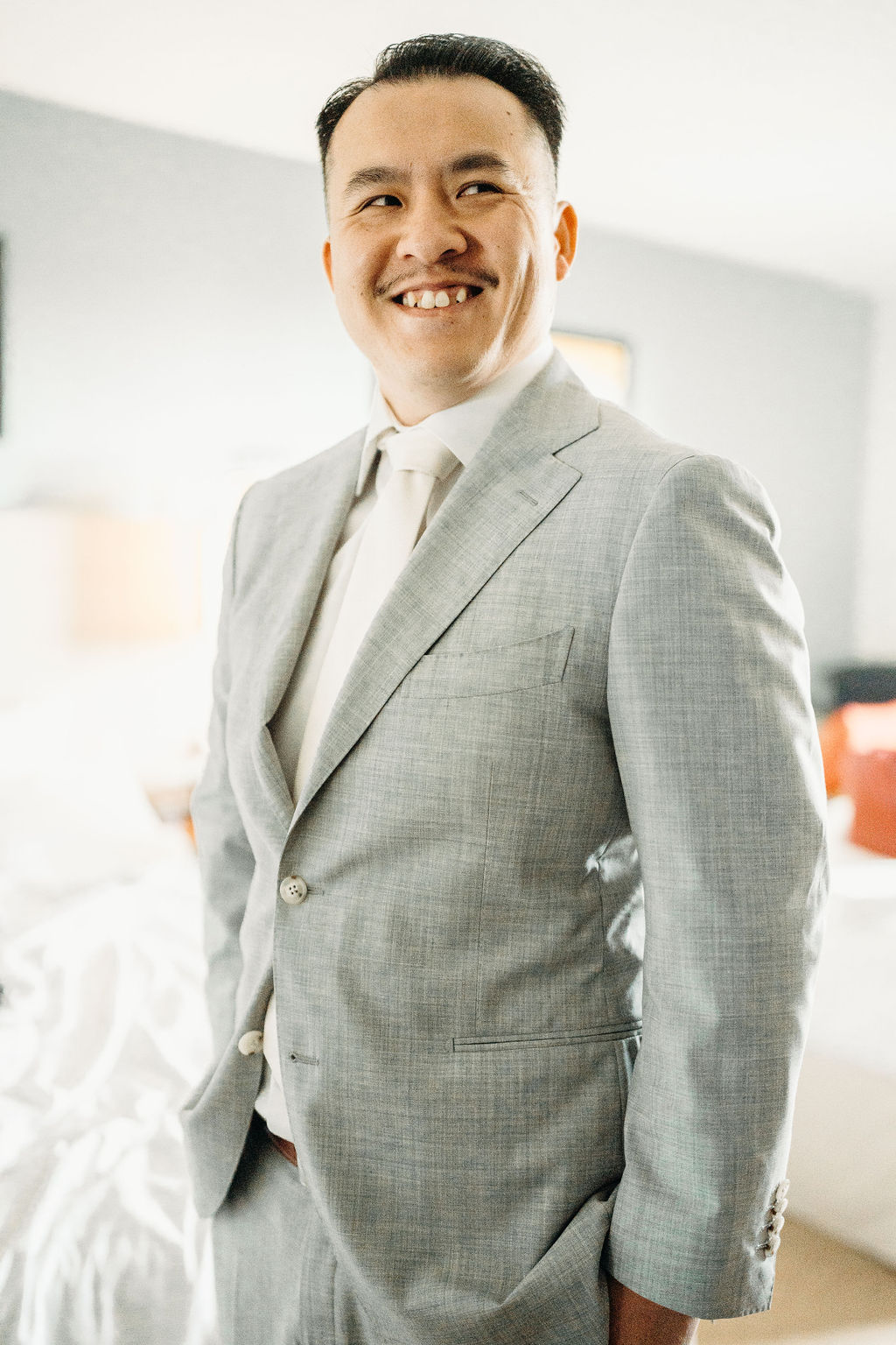 Man in a light gray suit stands smiling in a room with a bed and soft lighting.