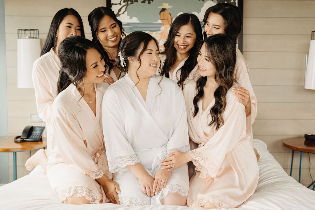 Seven women in robes sit and stand on a bed, smiling and interacting, with a light-colored wall and lamps in the background.