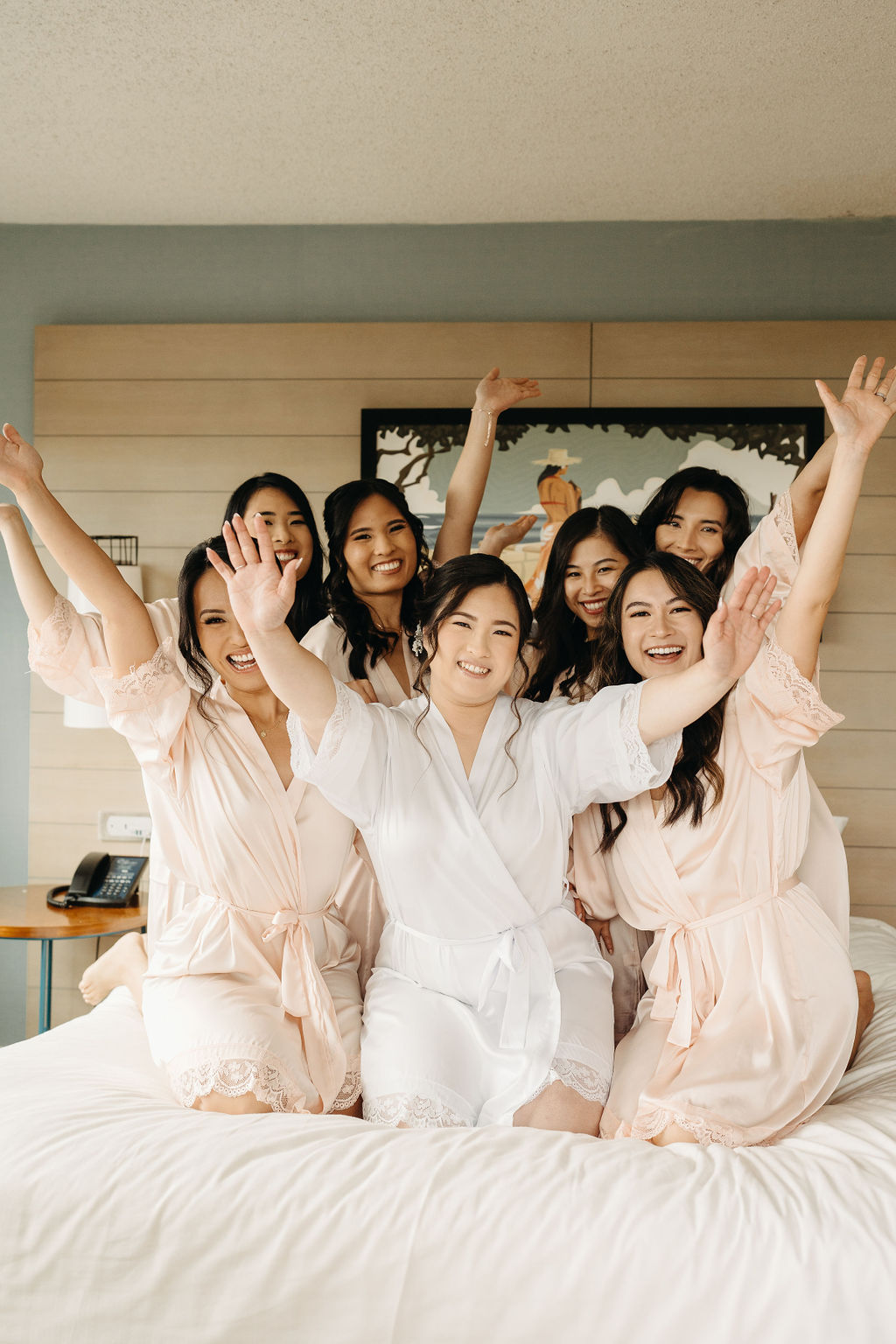 Seven women in robes sit and stand on a bed, smiling and interacting, with a light-colored wall and lamps in the background.