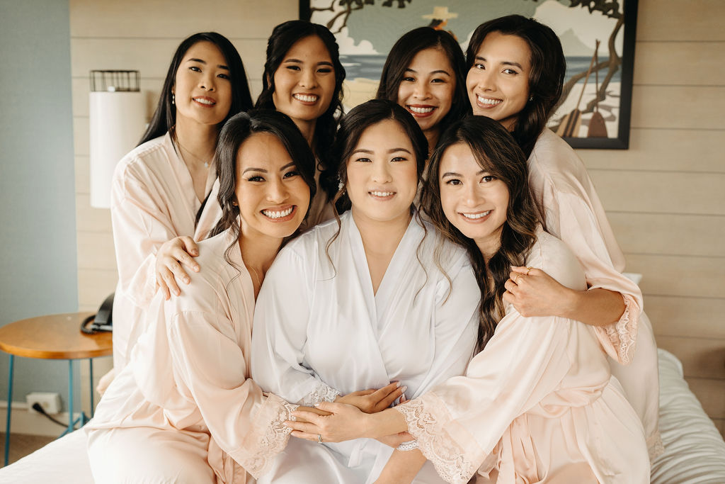 Seven women in robes sit and stand on a bed, smiling and interacting, with a light-colored wall and lamps in the background.