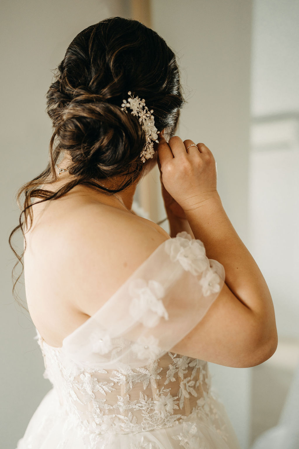 A bride adjusts her earring, wearing an off-shoulder wedding dress with lace details and a decorative hairpiece in an elegant updo.