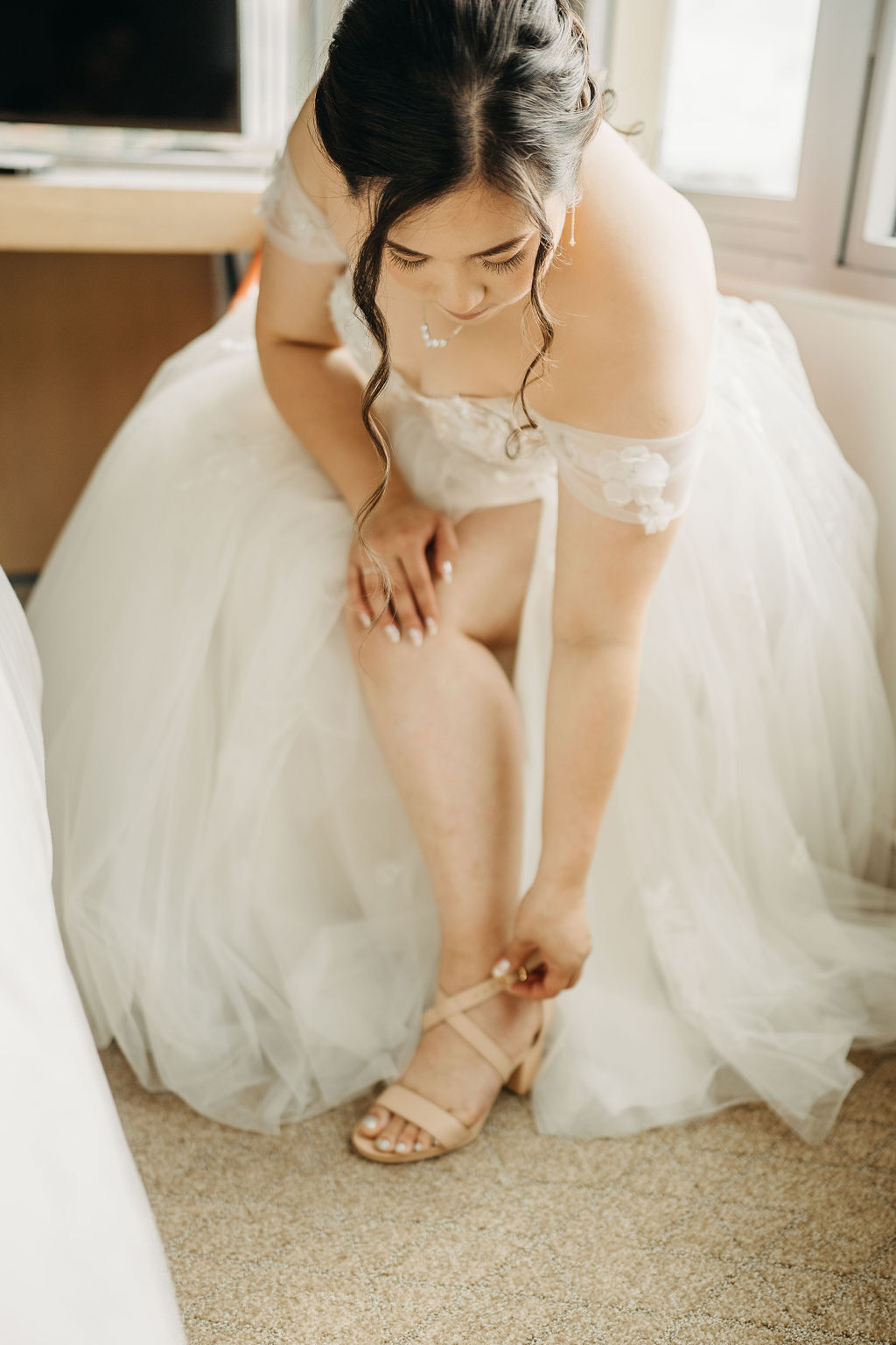 A bride adjusts her earring, wearing an off-shoulder wedding dress with lace details and a decorative hairpiece in an elegant updo.