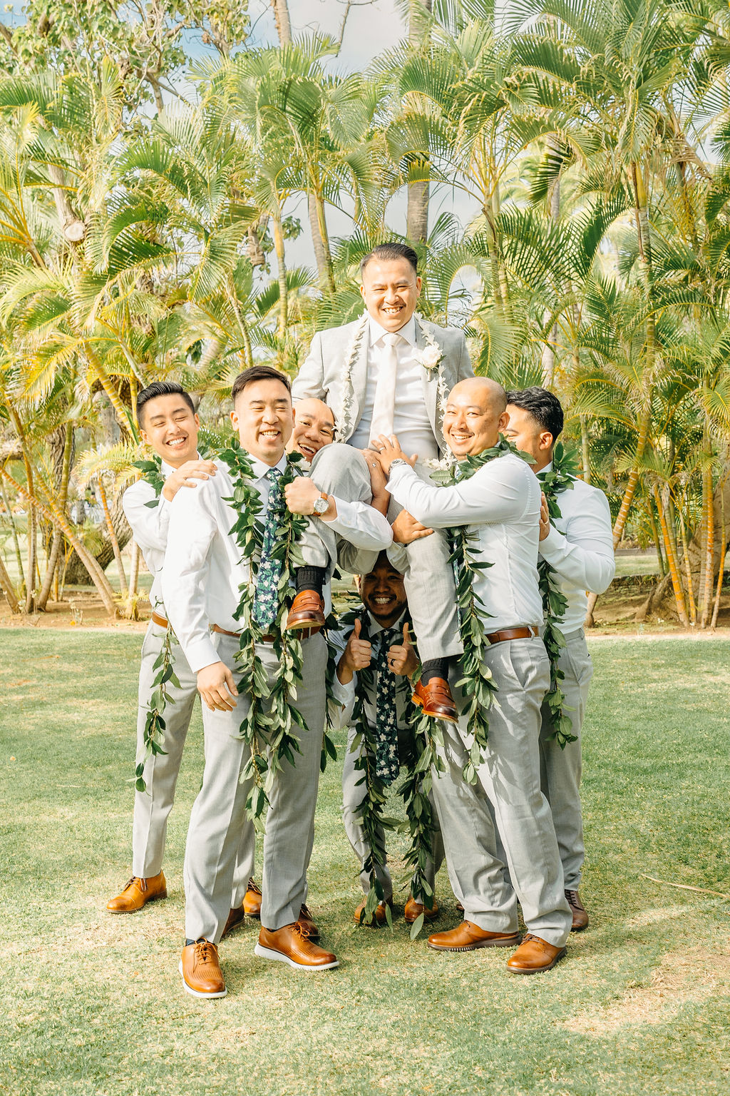 A group of groomsmen lifts the groom on their shoulders, smiling and wearing leis, posed in a sunny outdoor setting with palm trees for a wedding at Wedding at Lanikuhonua Cultural Institute