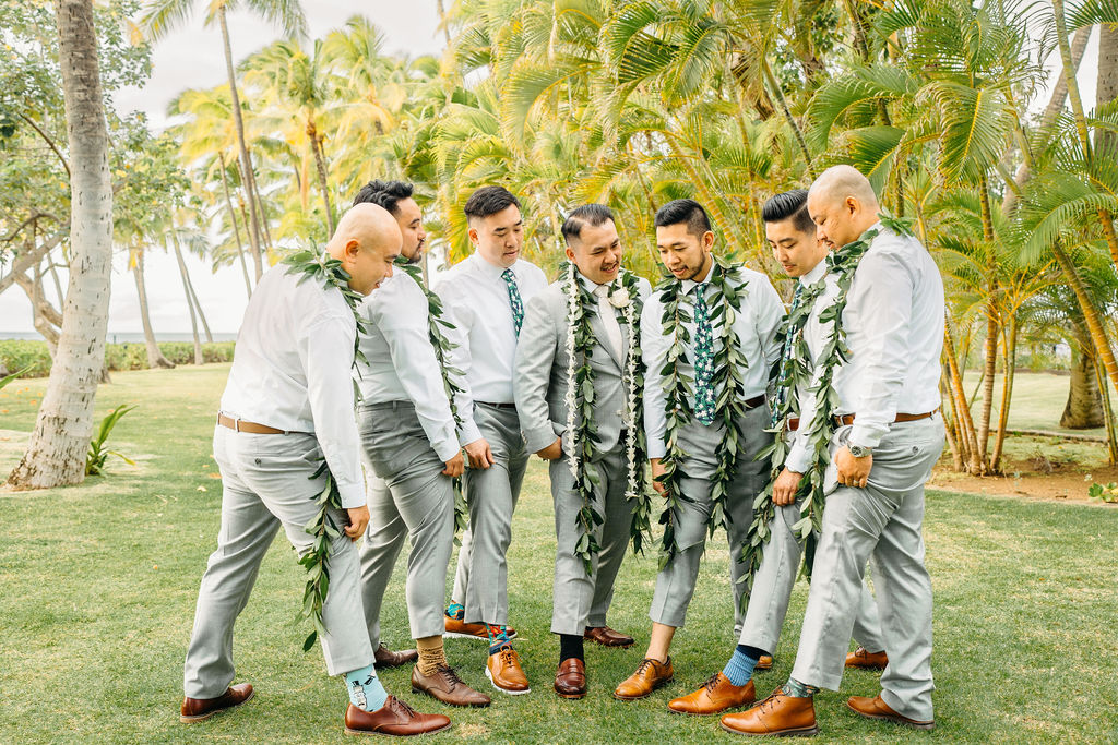 A group of groomsmen lifts the groom on their shoulders, smiling and wearing leis, posed in a sunny outdoor setting with palm trees for a wedding at Wedding at Lanikuhonua Cultural Institute