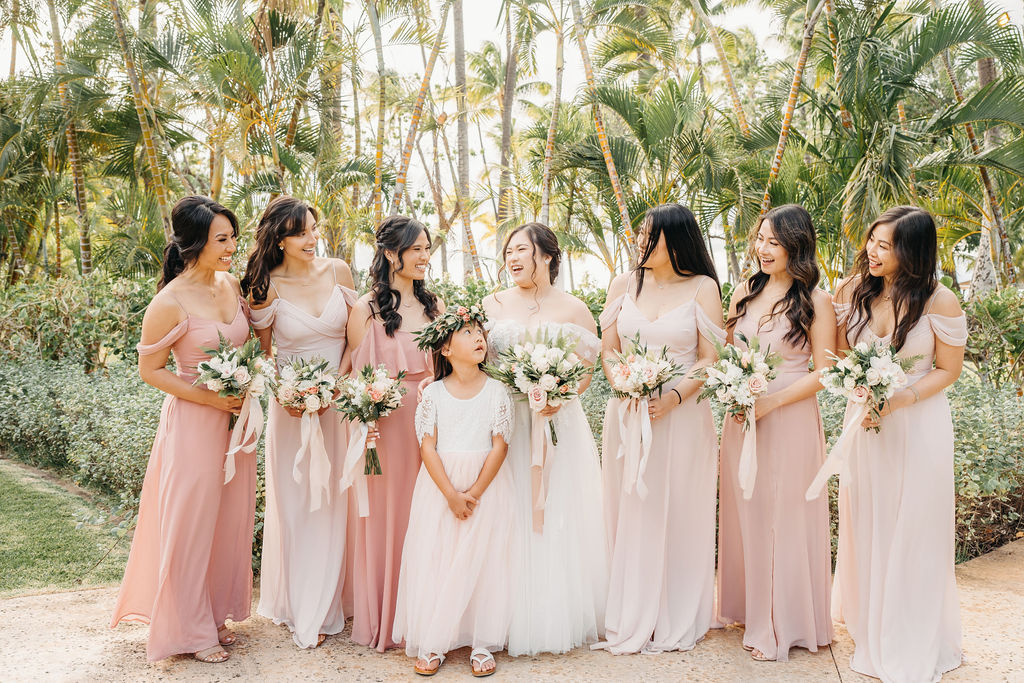 A bride in a white dress stands with six bridesmaids in pink dresses, holding bouquets, surrounded by palm trees for a Wedding at Lanikuhonua Cultural Institute