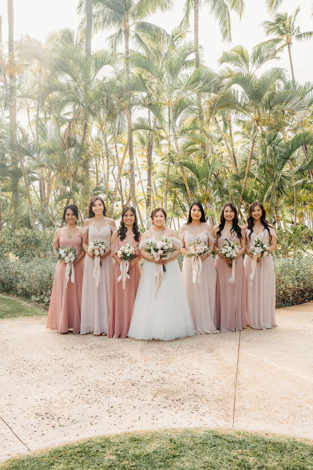 A bride in a white dress stands with six bridesmaids in pink dresses, holding bouquets, surrounded by palm trees for a Wedding at Lanikuhonua Cultural Institute