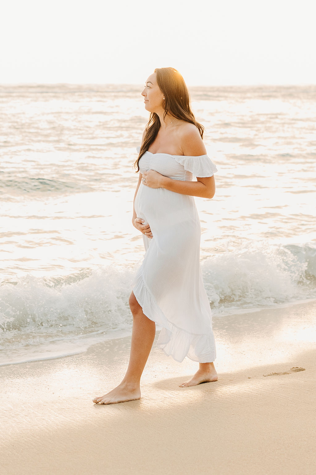 A woman in a white dress walks barefoot along a beach, holding her pregnant belly, with waves in the background.