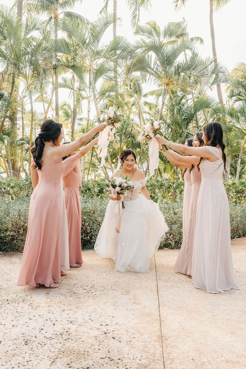 A bride in a white dress stands with six bridesmaids in pink dresses, holding bouquets, surrounded by palm trees for a Wedding at Lanikuhonua Cultural Institute