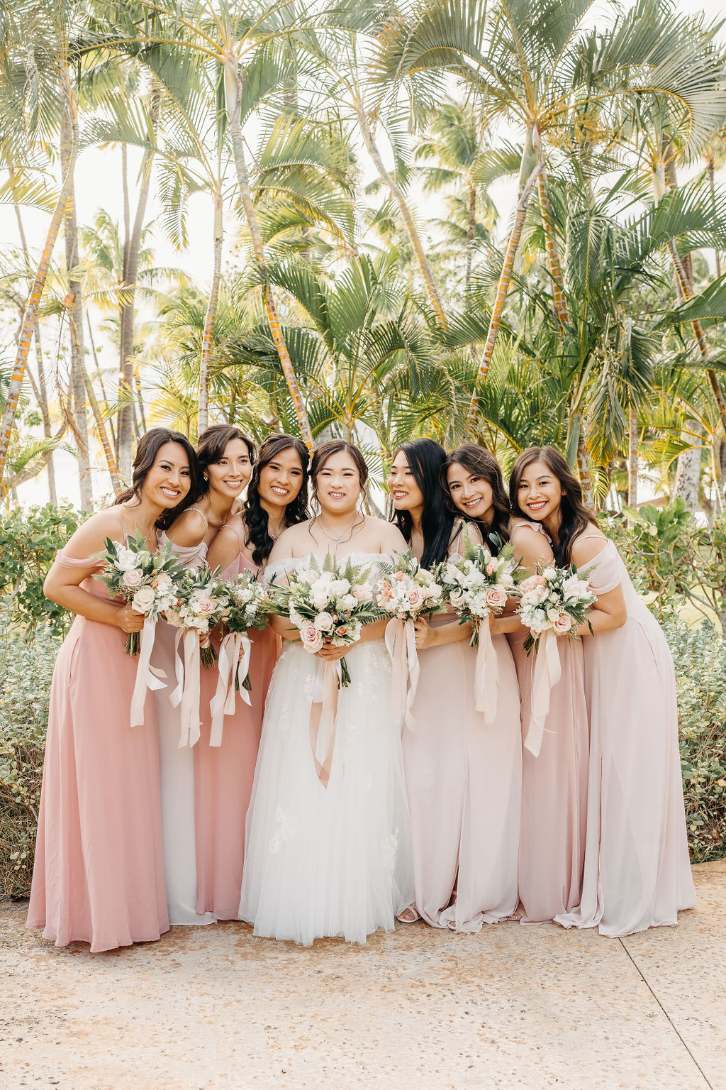 A bride in a white dress stands with six bridesmaids in pink dresses, holding bouquets, surrounded by palm trees for a Wedding at Lanikuhonua Cultural Institute