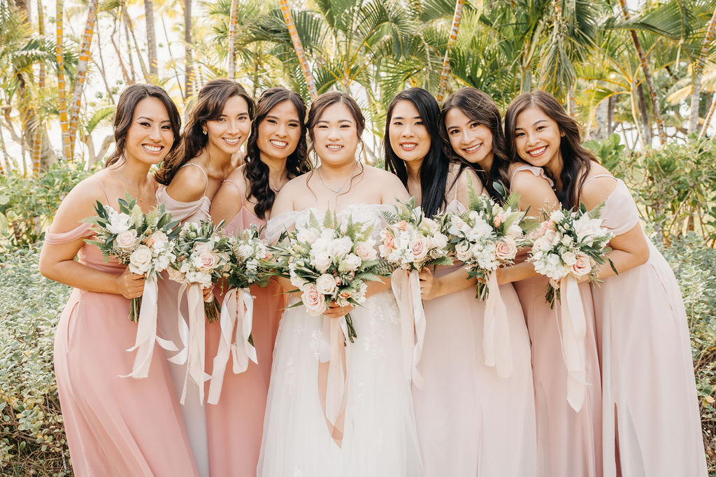 A bride in a white dress stands with six bridesmaids in pink dresses, holding bouquets, surrounded by palm trees for a Wedding at Lanikuhonua Cultural Institute