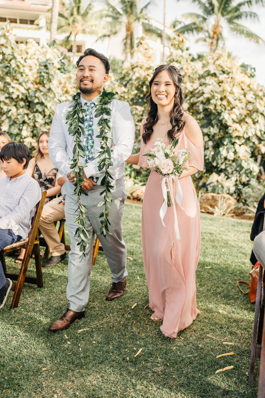 A man and woman walk down an outdoor aisle. The man wears a suit with a leafy garland, and the woman wears a pink dress holding a bouquet. Guests are seated on either side.