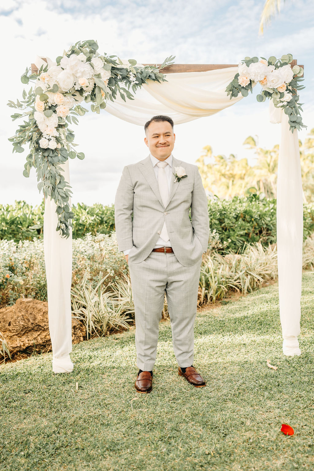 Person in a light gray suit stands under a flower-adorned wedding arch outdoors, with greenery and a cloudy sky in the background.