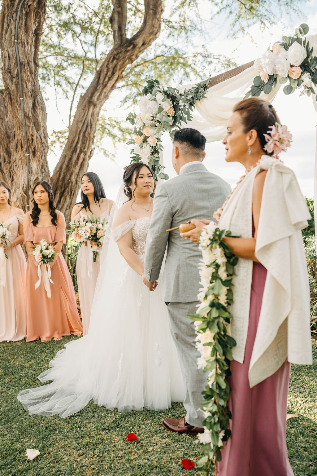 Bride and groom holding hands during an outdoor wedding ceremony. Bridesmaids in pink dresses stand nearby. A woman in a purple dress holds a floral garland for a Wedding at Lanikuhonua Cultural Institute