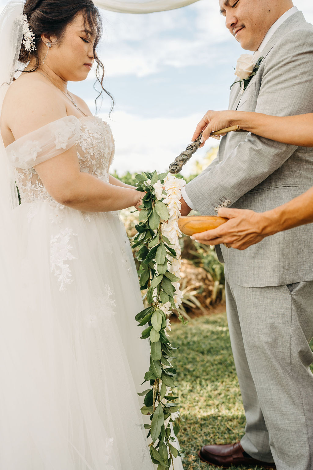 Bride and groom holding hands during an outdoor wedding ceremony. Bridesmaids in pink dresses stand nearby. A woman in a purple dress holds a floral garland for a Wedding at Lanikuhonua Cultural Institute