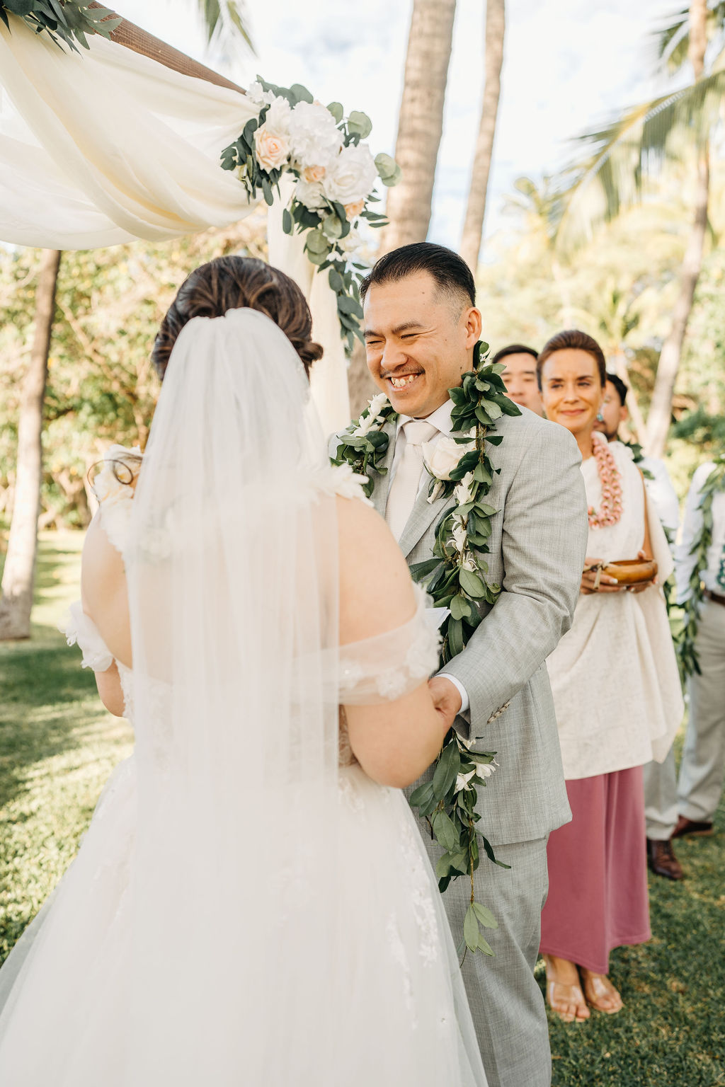 Bride and groom holding hands during an outdoor wedding ceremony. Bridesmaids in pink dresses stand nearby. A woman in a purple dress holds a floral garland for a Wedding at Lanikuhonua Cultural Institute