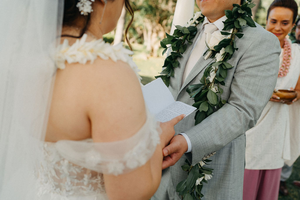 Bride and groom holding hands during an outdoor wedding ceremony. Bridesmaids in pink dresses stand nearby. A woman in a purple dress holds a floral garland for a Wedding at Lanikuhonua Cultural Institute