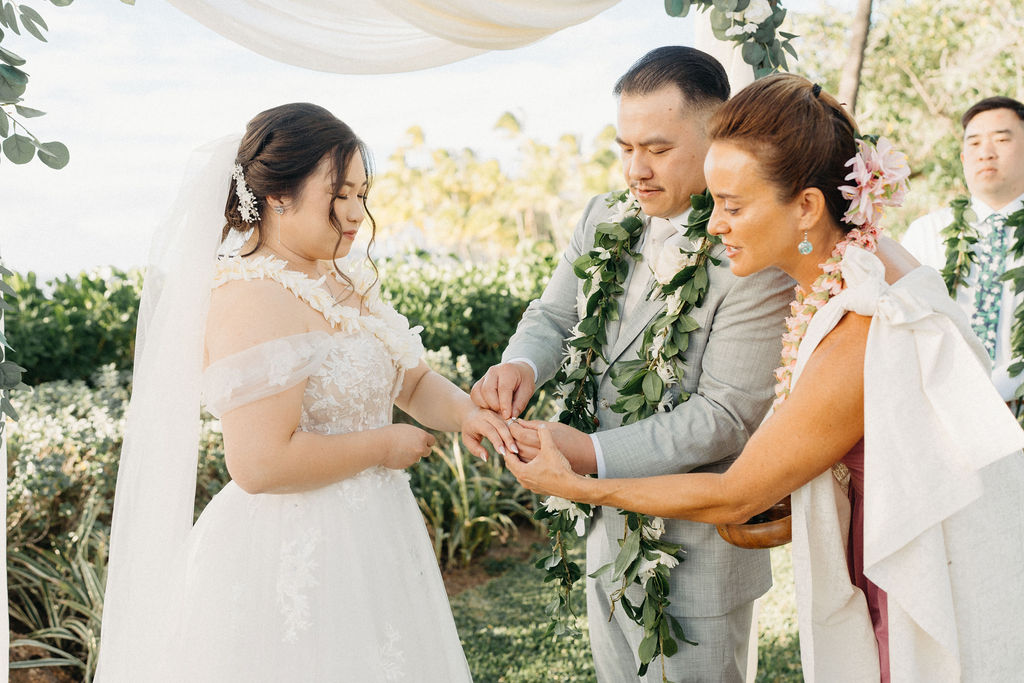 Bride and groom holding hands during an outdoor wedding ceremony. Bridesmaids in pink dresses stand nearby. A woman in a purple dress holds a floral garland for a Wedding at Lanikuhonua Cultural Institute