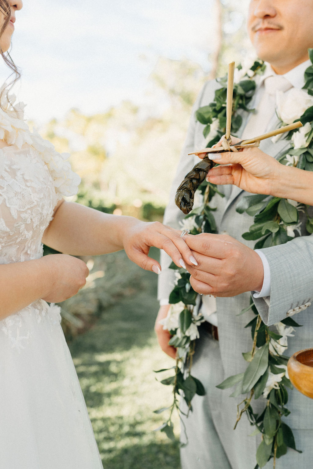 Bride and groom holding hands during an outdoor wedding ceremony. Bridesmaids in pink dresses stand nearby. A woman in a purple dress holds a floral garland for a Wedding at Lanikuhonua Cultural Institute