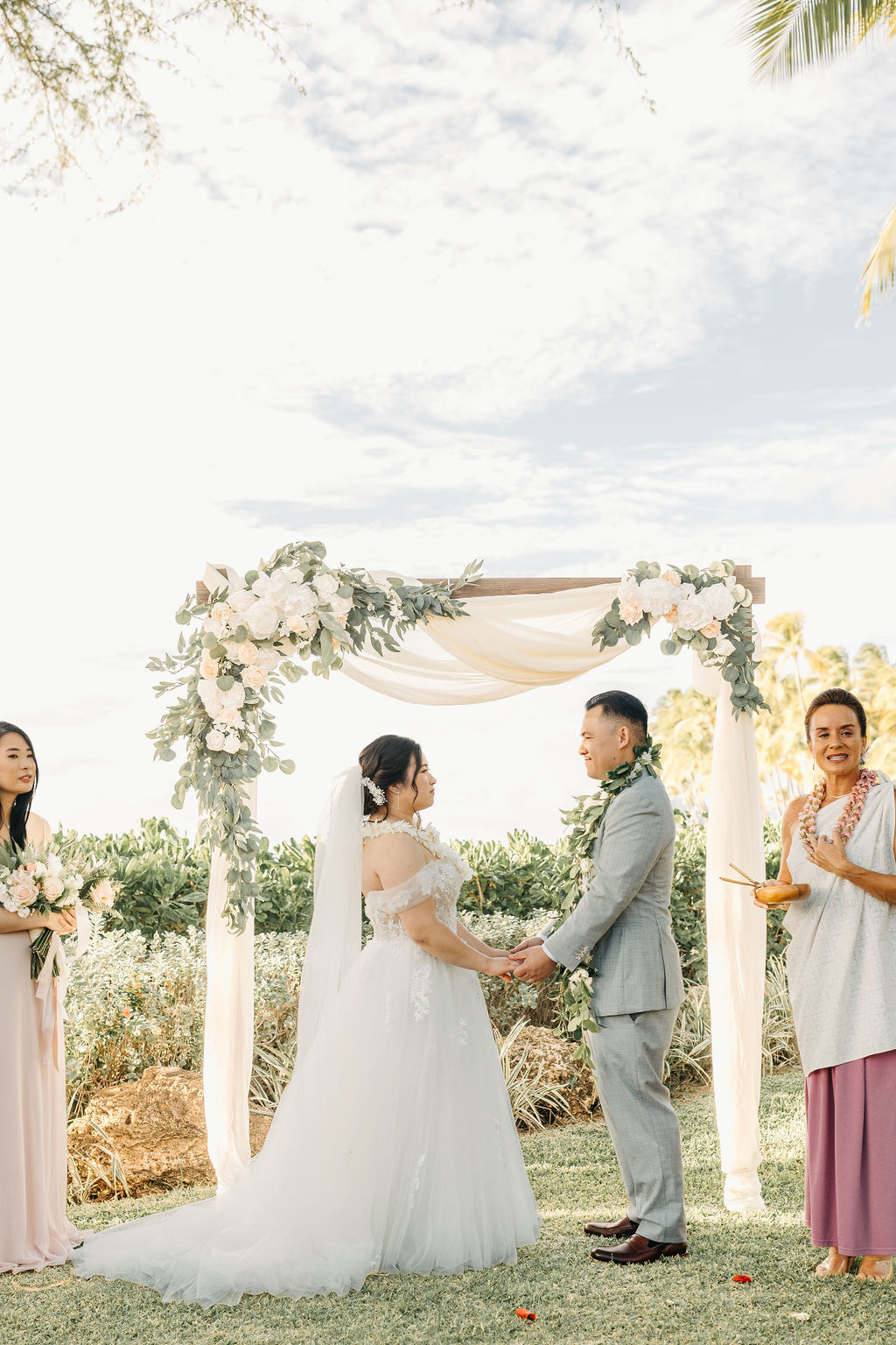 Bride and groom holding hands during an outdoor wedding ceremony. Bridesmaids in pink dresses stand nearby. A woman in a purple dress holds a floral garland for a Wedding at Lanikuhonua Cultural Institute