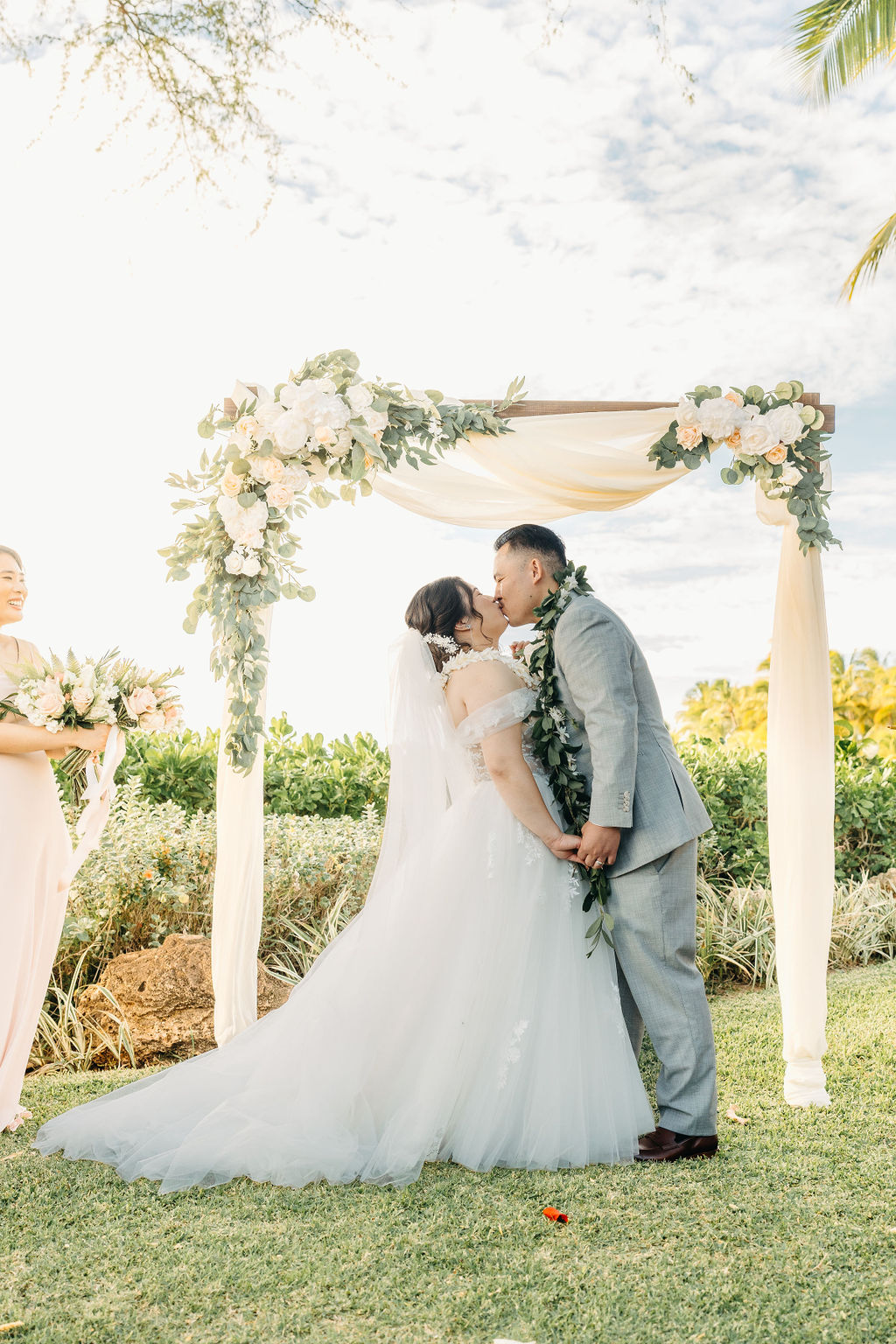 Bride and groom holding hands during an outdoor wedding ceremony. Bridesmaids in pink dresses stand nearby. A woman in a purple dress holds a floral garland for a Wedding at Lanikuhonua Cultural Institute