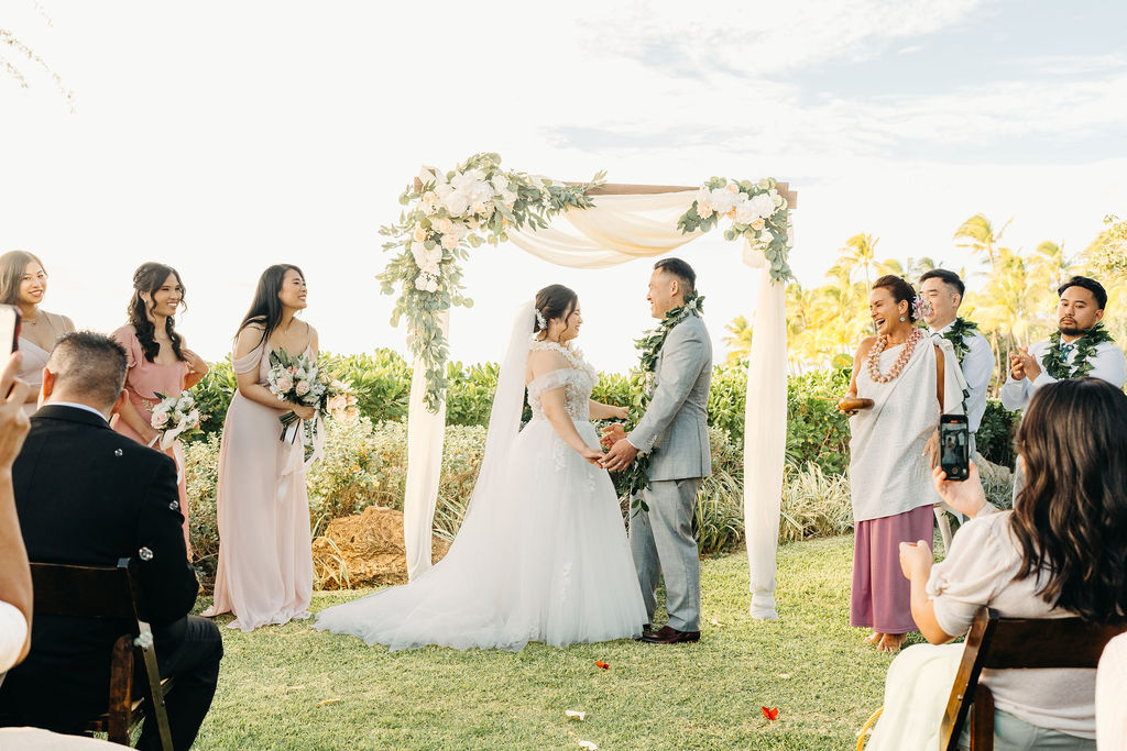 Bride and groom holding hands during an outdoor wedding ceremony. Bridesmaids in pink dresses stand nearby. A woman in a purple dress holds a floral garland for a Wedding at Lanikuhonua Cultural Institute