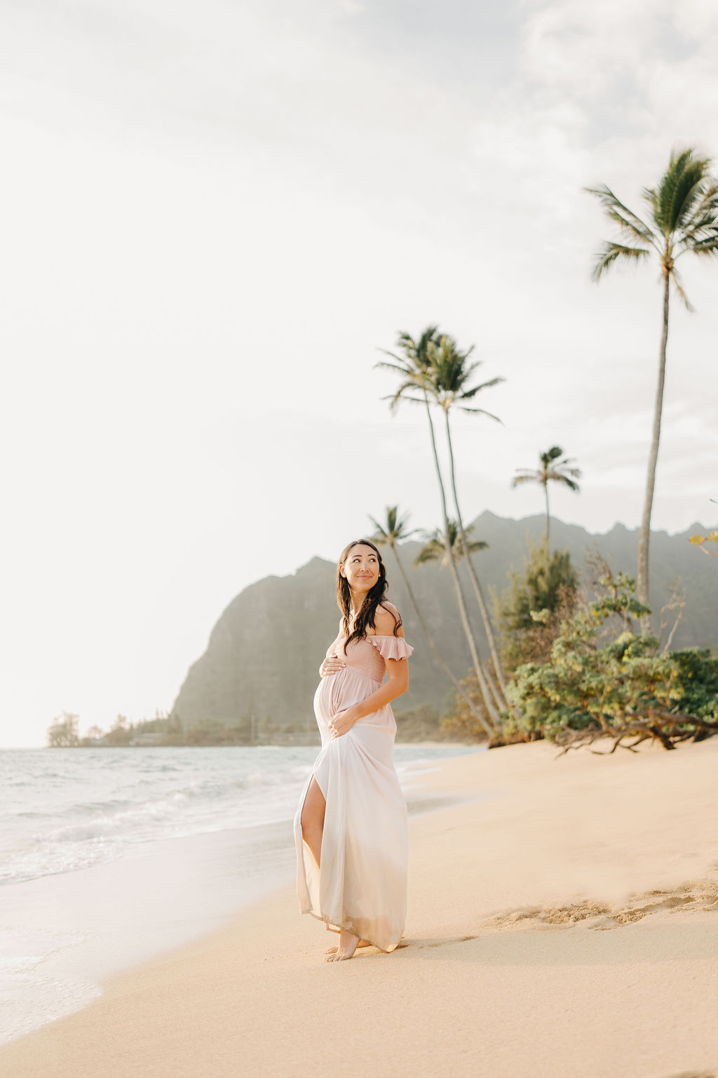 A woman in a white dress walks barefoot along a beach, holding her pregnant belly, with waves in the background.