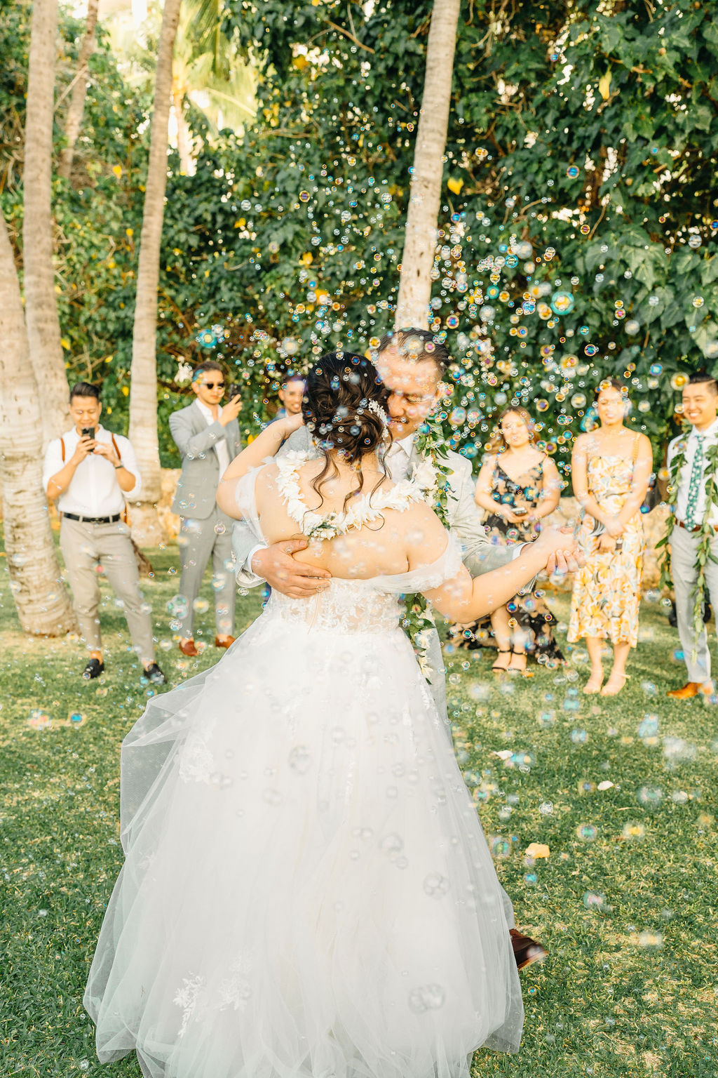 A couple dances in a garden, surrounded by bubbles. The groom wears a gray suit with a floral garland, and the bride wears a white gown with lace detailing.