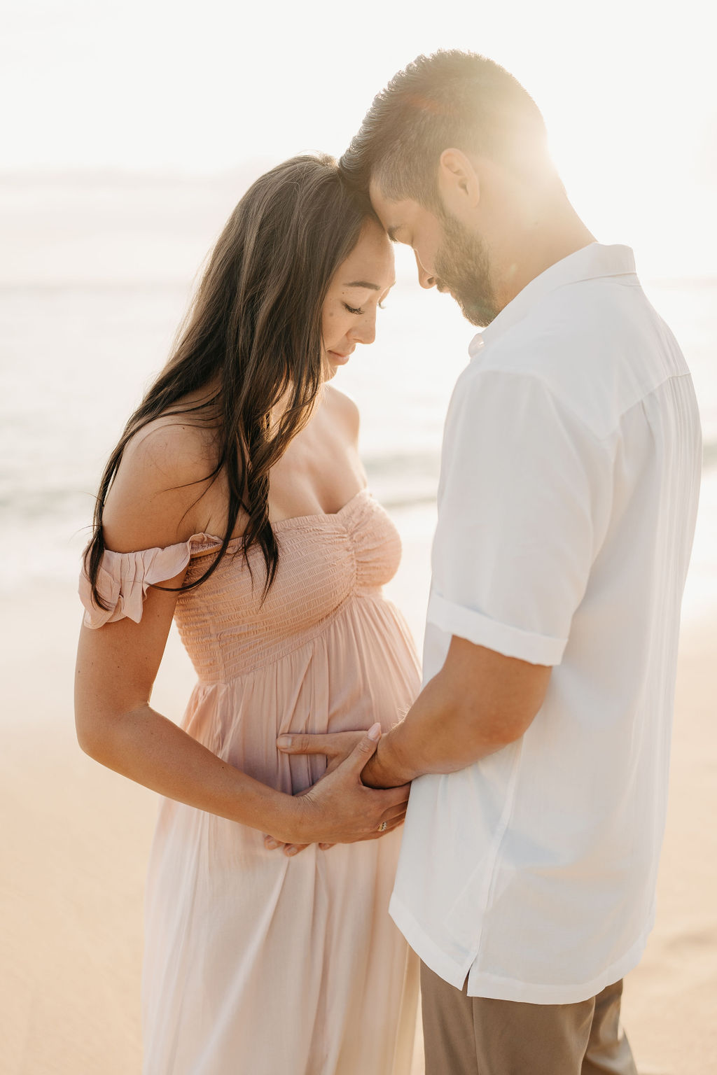 A woman in a white dress walks barefoot along a beach, holding her pregnant belly, with waves in the background.