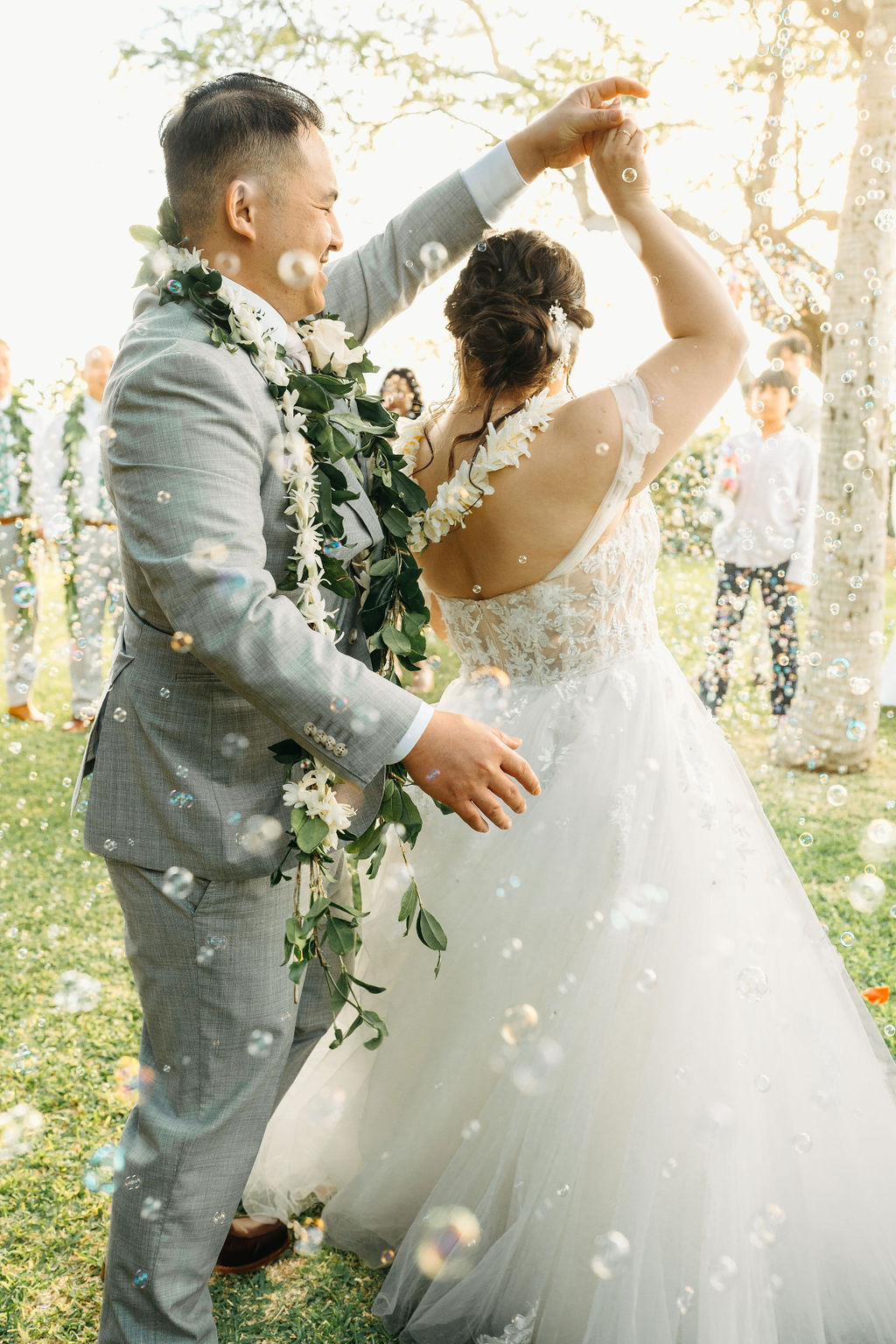 A couple dances in a garden, surrounded by bubbles. The groom wears a gray suit with a floral garland, and the bride wears a white gown with lace detailing.