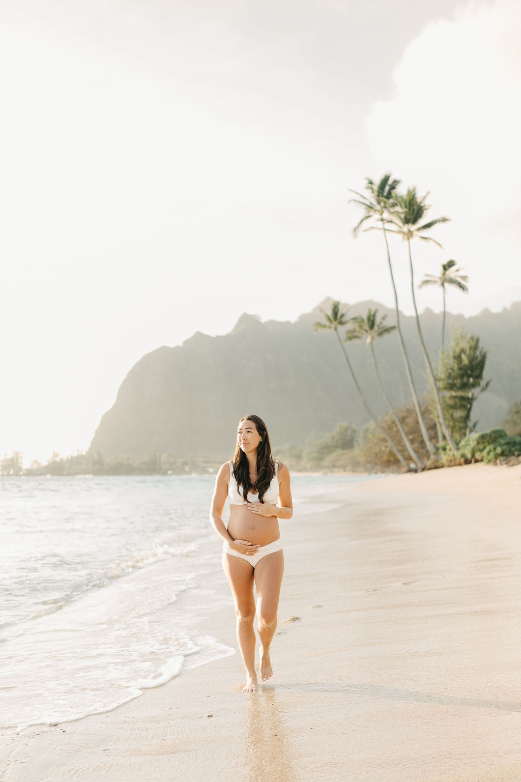 A woman in a white bikini walks along a sandy beach with gentle waves. There are palm trees and distant cliffs in the background under a bright sky for a beach maternity session