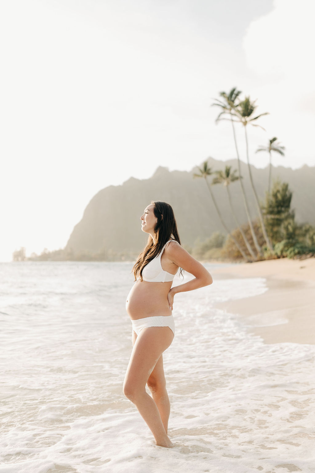 A woman in a white bikini walks along a sandy beach with gentle waves. There are palm trees and distant cliffs in the background under a bright sky for a beach maternity session