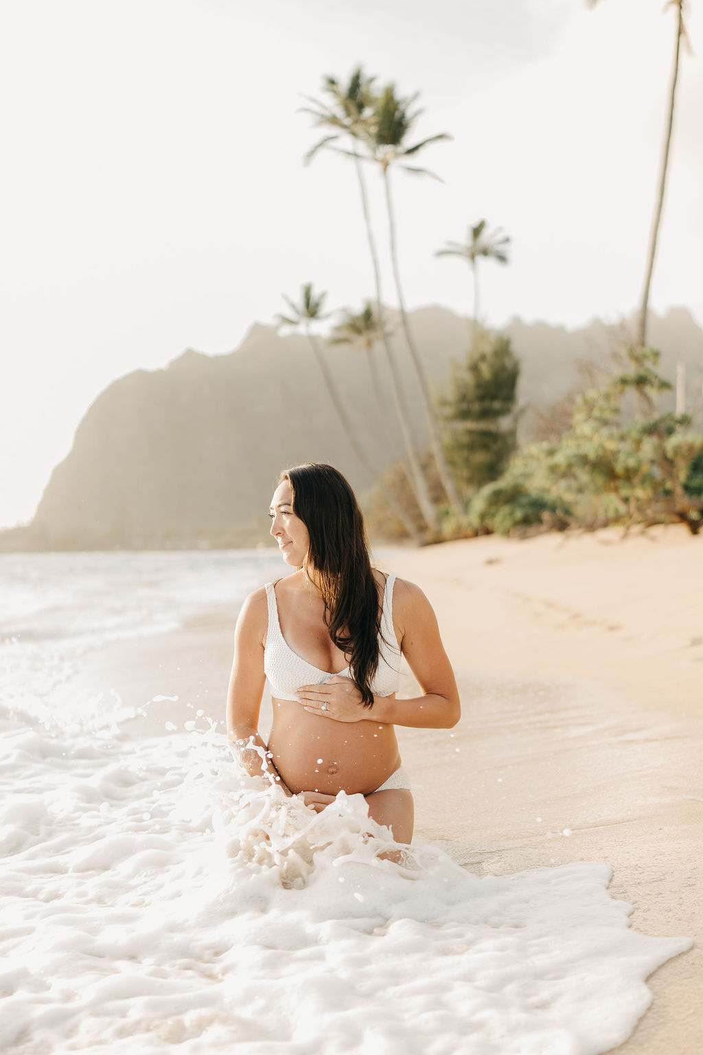 A woman in a white bikini walks along a sandy beach with gentle waves. There are palm trees and distant cliffs in the background under a bright sky for a beach maternity session