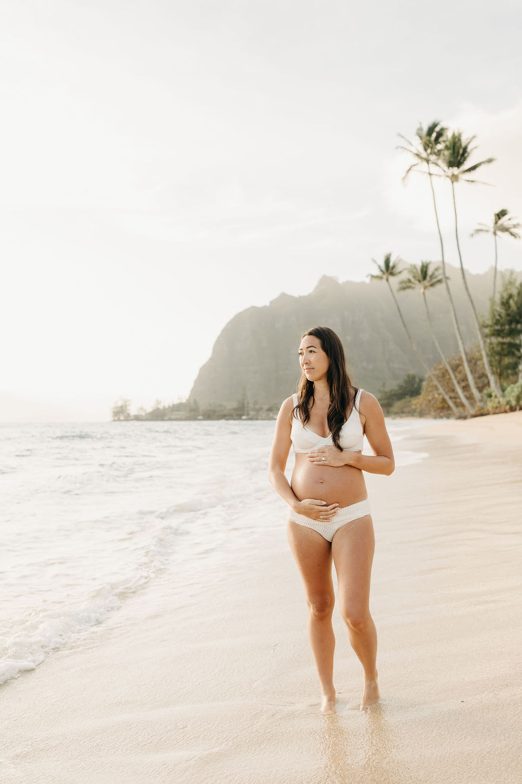 Pregnant woman in a white bikini stands on a sandy beach, touching her belly, with palm trees and mountains in the background.