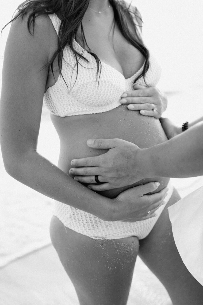 Pregnant woman in a white bikini stands on a sandy beach, touching her belly, with palm trees and mountains in the background.