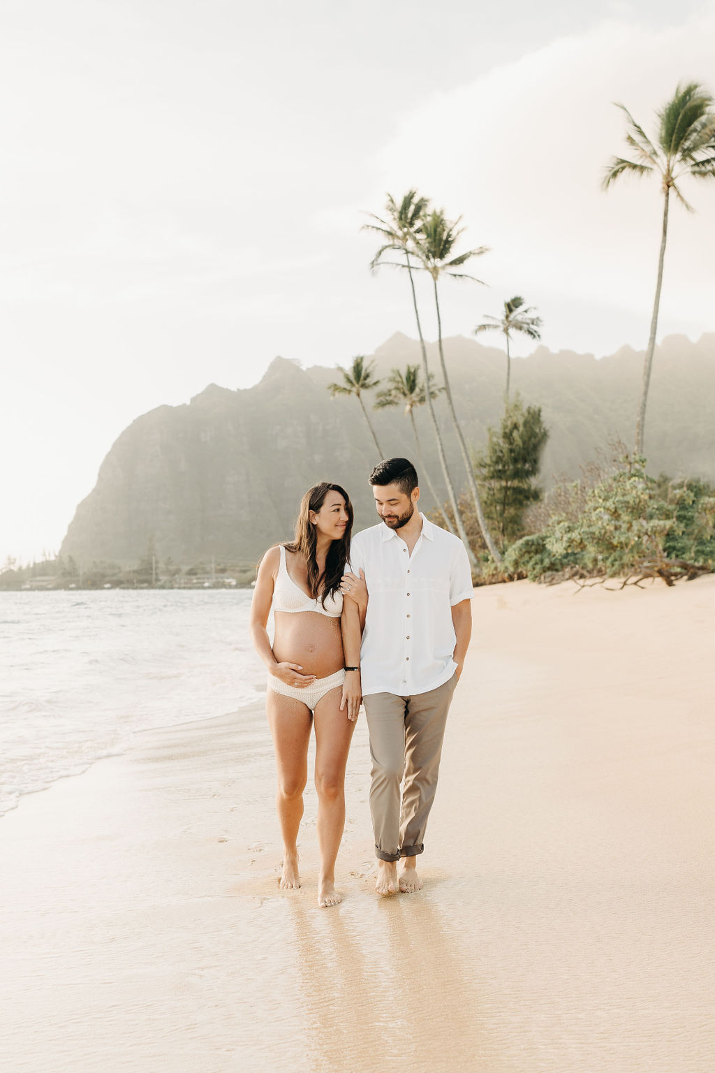 A pregnant woman and a man walk barefoot on a sandy beach, with palm trees and mountains in the background.