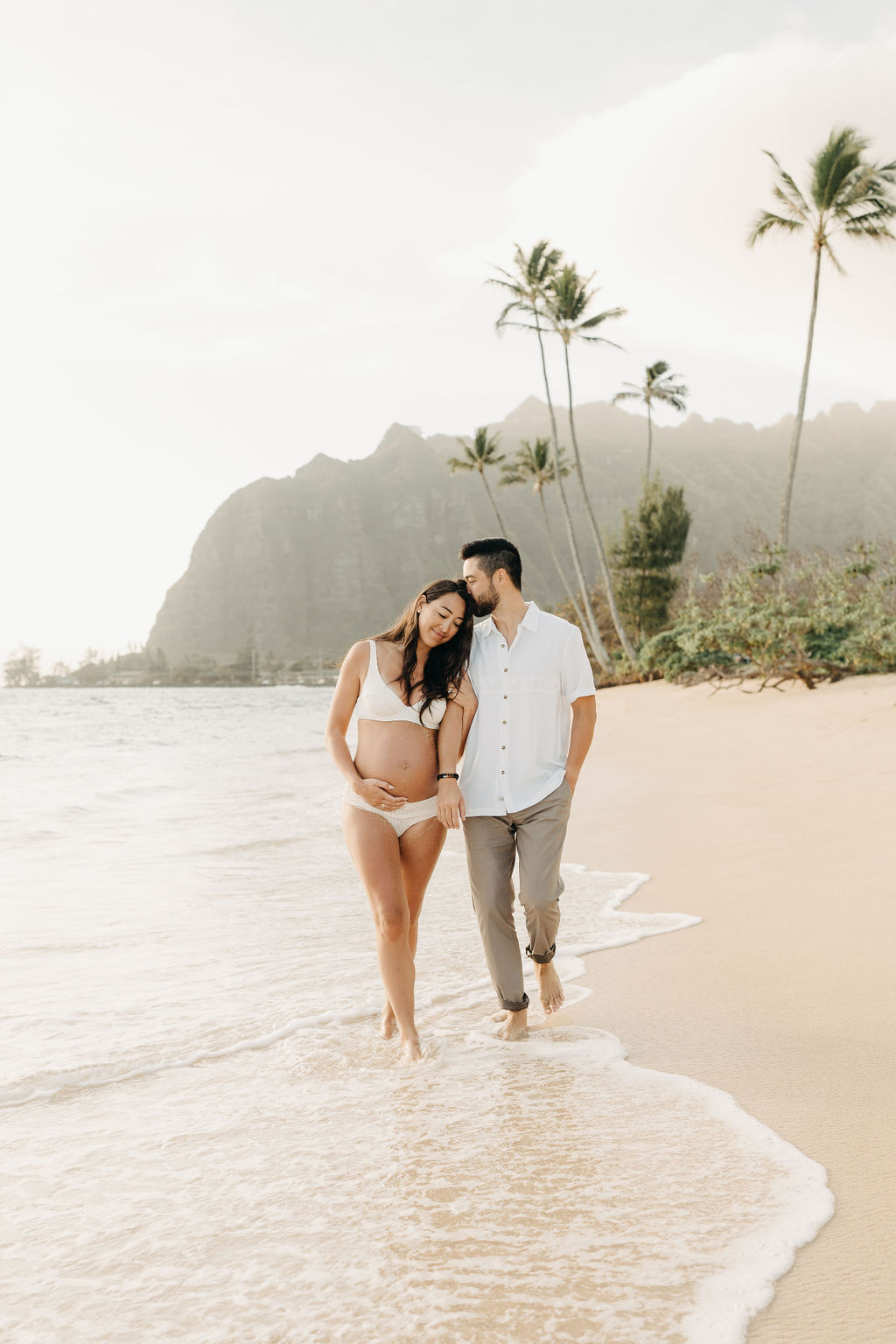 A pregnant woman and a man walk barefoot on a sandy beach, with palm trees and mountains in the background.