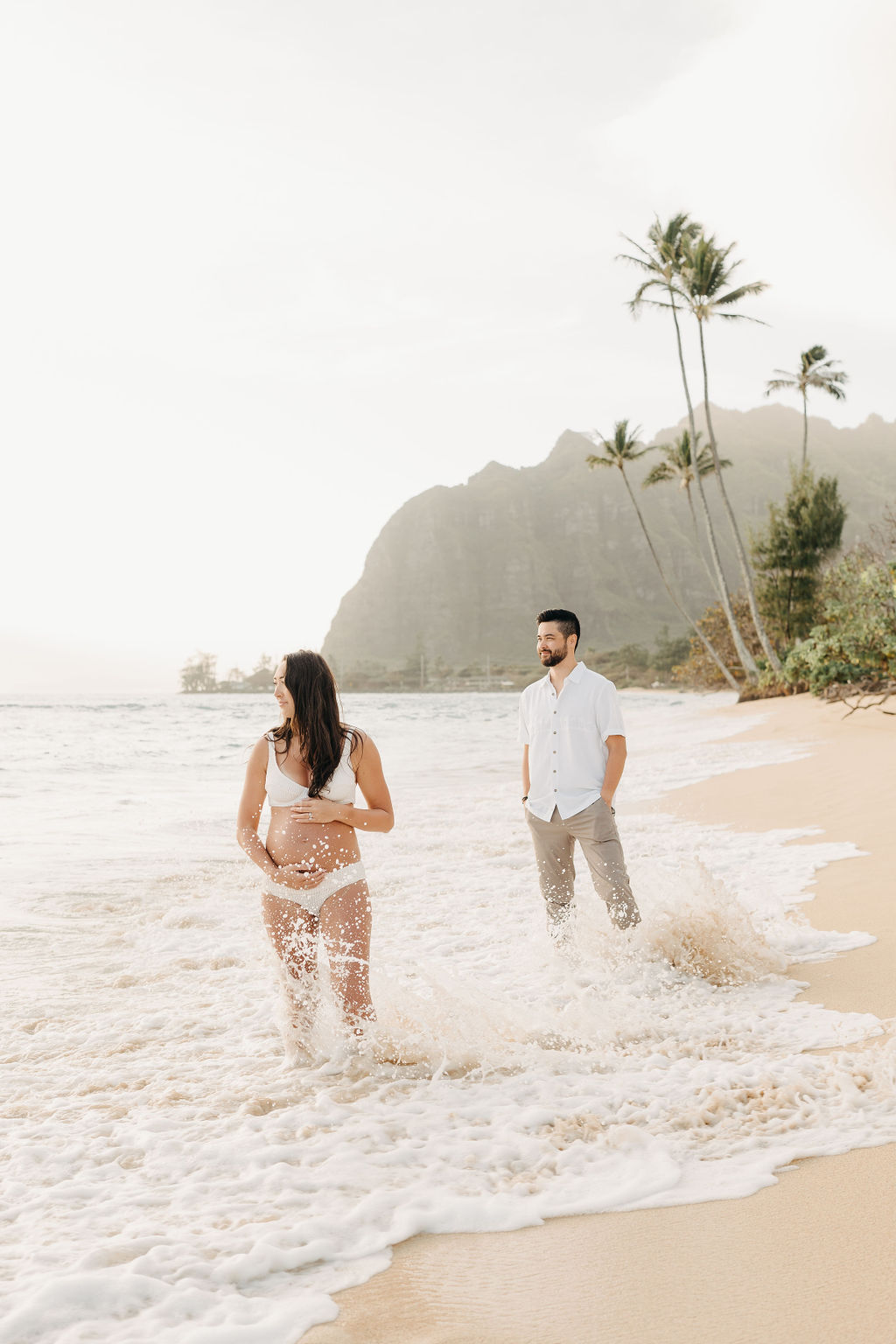 A pregnant woman and a man walk barefoot on a sandy beach, with palm trees and mountains in the background.