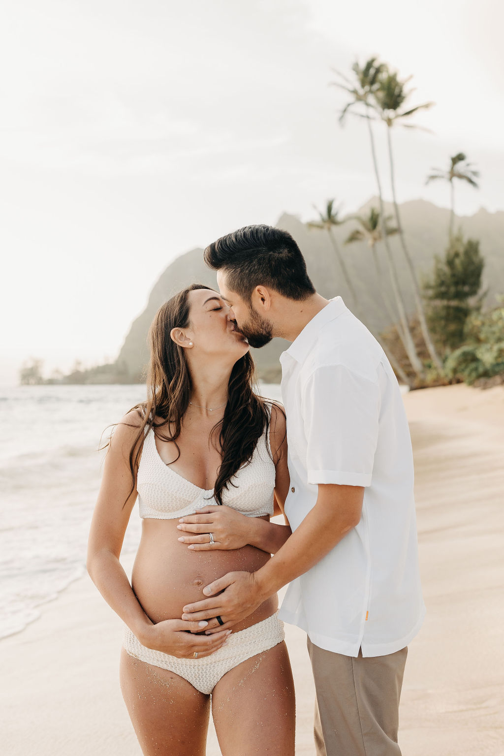 A pregnant woman and a man walk barefoot on a sandy beach, with palm trees and mountains in the background.