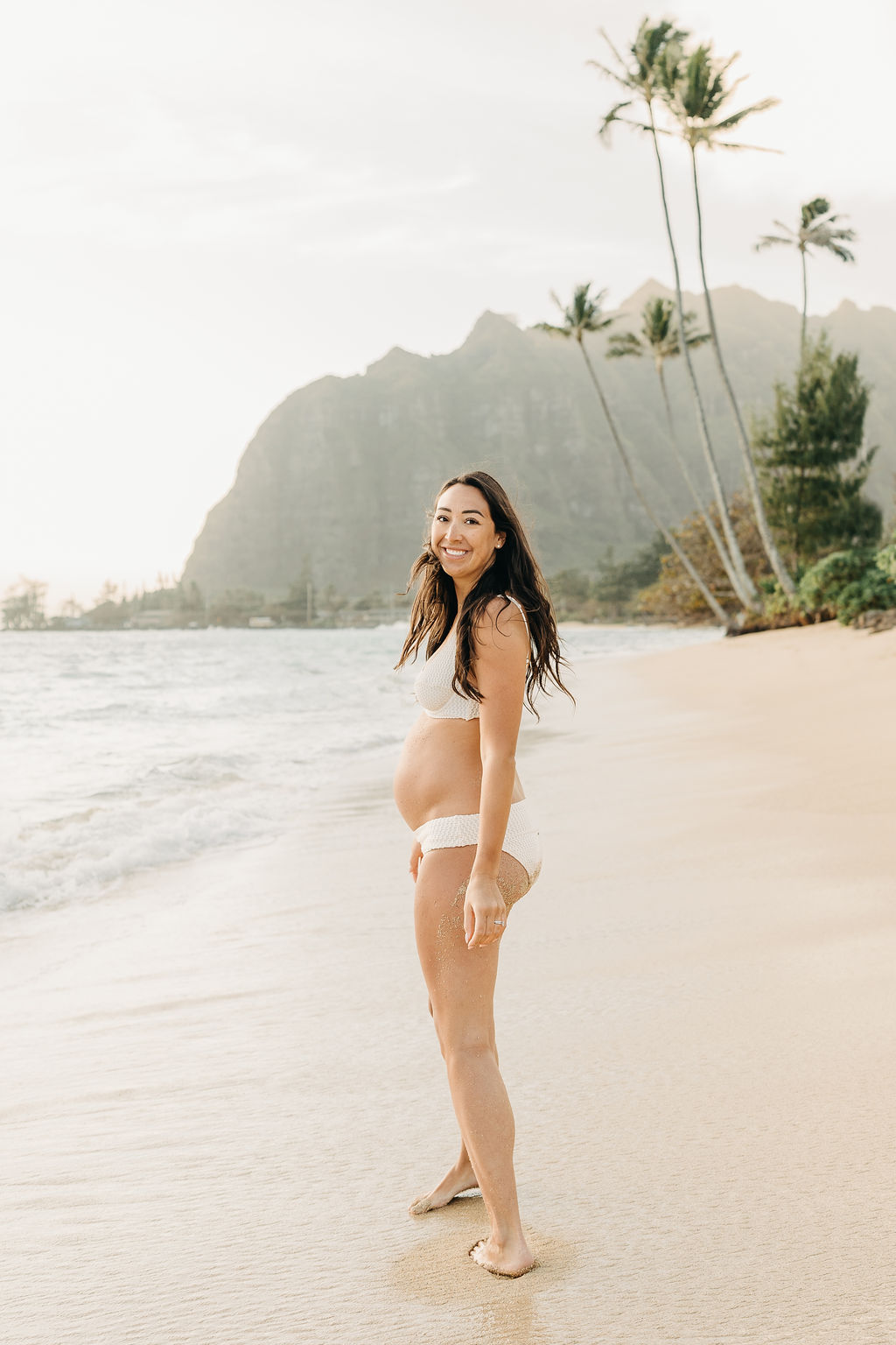 A pregnant woman and a man walk barefoot on a sandy beach, with palm trees and mountains in the background.