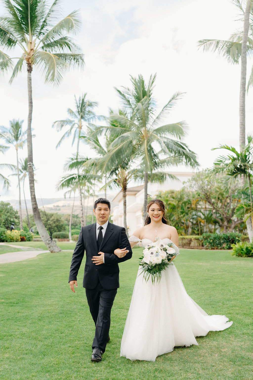 A bride in a white gown holds a bouquet and walks arm-in-arm with a man in a dark suit on a grassy path lined with palm trees.