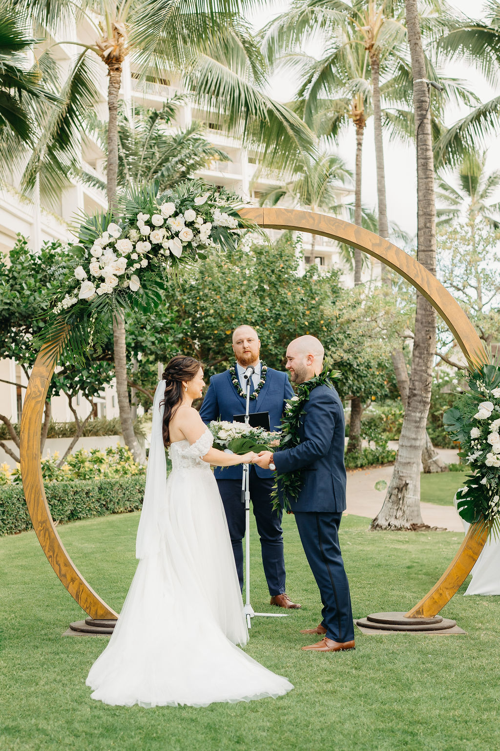 A bride and groom stand facing each other holding hands under a circular floral arch, with an officiant in the background. Palm trees and greenery surround the scene for a wedding at the four seasons in oahu